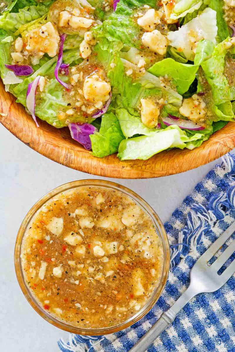 n overhead view of a bowl of Outback blue cheese vinaigrette next to a large wooden salad bowl. The fresh greens in the salad bowl are tossed with the chunky, herb-flecked vinaigrette, ready to be served.