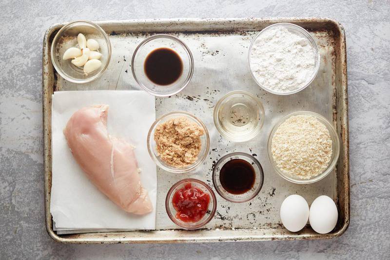An overhead view of the ingredients for chicken katsu and tonkatsu sauce arranged on a baking sheet. Pictured are a raw chicken breast, eggs, and bowls of flour, panko, soy sauce, mirin, ketchup, brown sugar, and garlic.