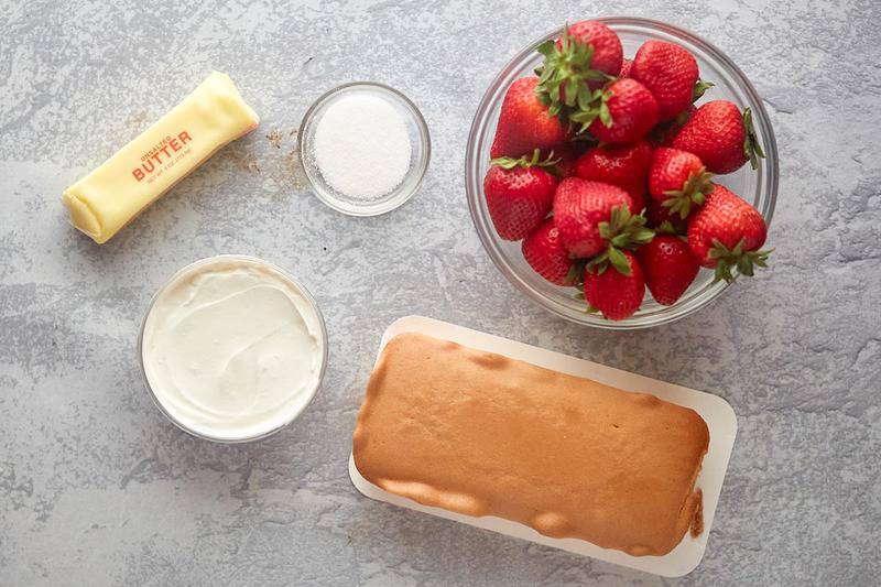 Overhead view of ingredients: fresh strawberries, vanilla yogurt, whipped cream, butter, sugar, and pound cake cubes on a tray.
