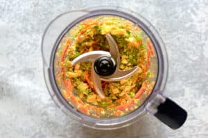 A top-down view into a food processor, showing a colorful mix of cooked vegetables like carrots, corn, and peas that have been finely chopped for the soup.