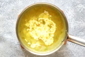 A rustic brown soup crock filled with creamy vegetable soup, garnished with a colorful mix of whole vegetables. A spoon rests beside the bowl on a striped napkin.