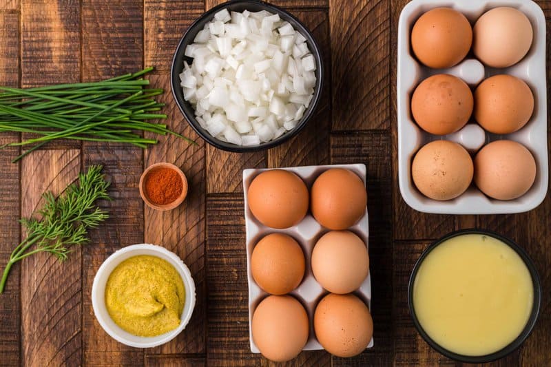 An overhead view of the ingredients for smoked deviled eggs on a wooden surface. Pictured are cartons of eggs, bowls of diced onion, slaw dressing, and mustard, along with fresh chives, dill, and a small dish of paprika.