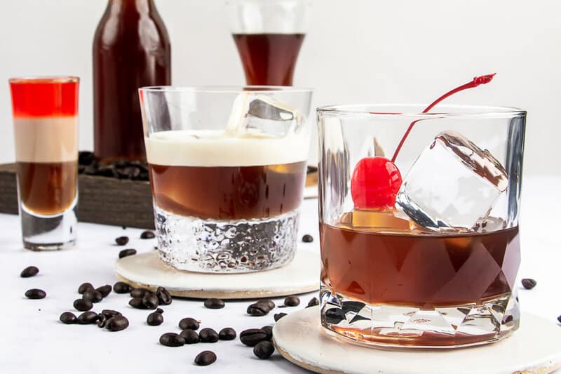 Several cocktails made with homemade Kahlua are displayed, including a White Russian, a Black Russian with a cherry, and a layered B-52 shot. A bottle of the coffee liqueur sits in the background.