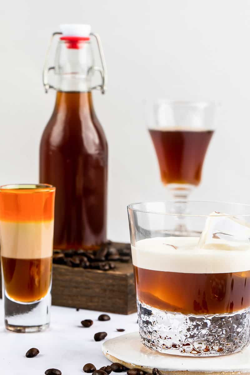 A close-up of different cocktails made with homemade Kahlua. A White Russian with a cream float and a colorful, layered B-52 shot are in sharp focus, with the bottle of Kahlua in the background.