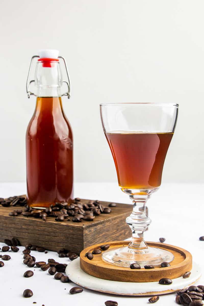 A bottle of homemade Kahlua is displayed next to a stemmed glass filled with the dark coffee liqueur, ready for sipping. The scene is decorated with scattered coffee beans.