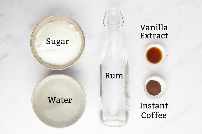 An overhead view of the labeled ingredients for homemade Kahlua. Pictured are bowls of sugar and water, dishes of vanilla extract and instant coffee, and an empty glass bottle labeled "Rum."