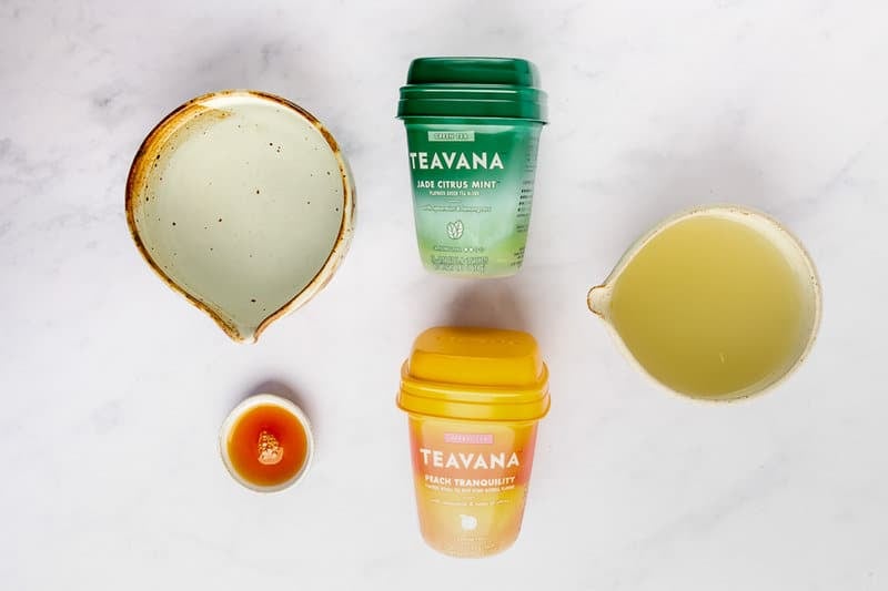 An overhead view of the ingredients for a Starbucks Medicine Ball tea. Pictured are containers of Teavana Jade Citrus Mint and Peach Tranquility teas, along with pitchers of lemonade and water, and a small dish of honey.