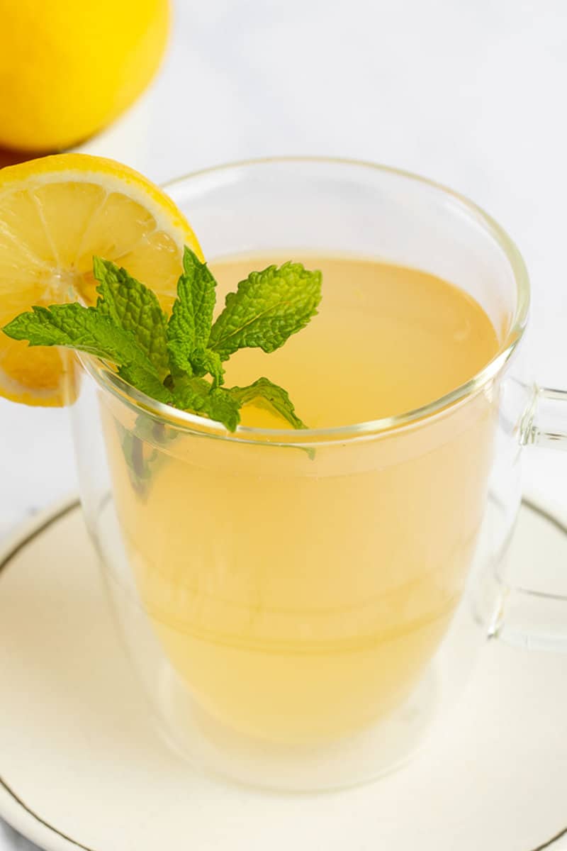 A close-up of a glass mug filled with warm, golden Medicine Ball tea. The comforting drink is garnished with a bright slice of lemon and a sprig of fresh mint.