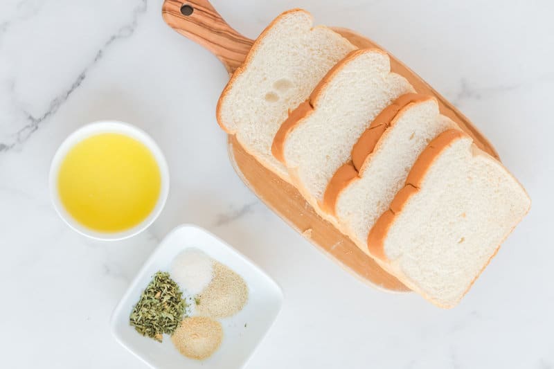An overhead view of the ingredients for air fryer croutons on a white marble surface. Pictured are several slices of white bread on a cutting board, a small bowl of melted butter, and a dish containing mounds of dried parsley, onion powder, garlic powder, and salt.