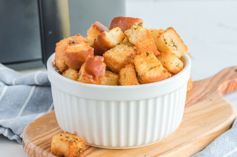 A close-up of a white bowl filled with a generous amount of golden-brown air fryer croutons. The image highlights the crunchy texture and the specks of herbs coating each piece.