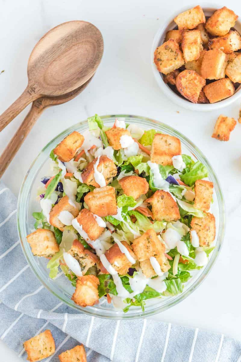 An overhead view of a large glass bowl of salad topped with crispy, golden-brown air fryer croutons and a drizzle of dressing. A smaller bowl of croutons and wooden salad servers are positioned next to the main bowl, showcasing a perfect serving suggestion.