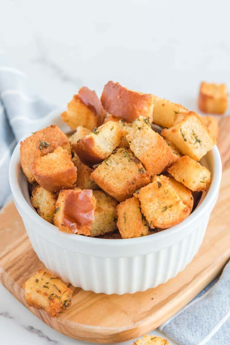 A white ceramic bowl is filled with crispy, golden-brown homemade croutons seasoned with herbs. The bowl sits on a wooden board, with the air fryer partially visible in the blurred background.