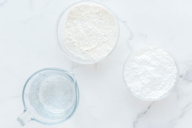An overhead view of the three simple ingredients for tempura batter on a white marble surface. Pictured are a glass bowl of all-purpose flour, a smaller bowl of cornstarch, and a measuring cup of club soda.