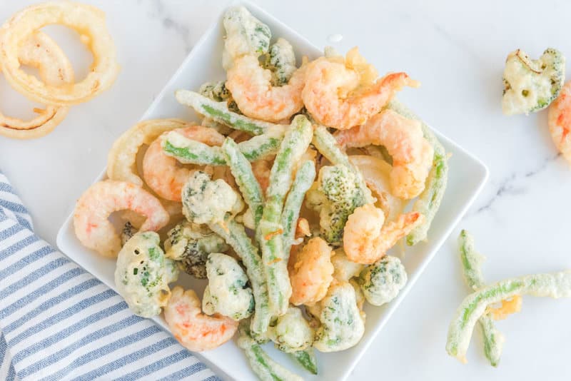A white square plate filled with assorted freshly made tempura. The light, crispy batter coats pink shrimp, green broccoli florets, green beans, and golden onion rings, showcasing the variety of foods that can be used.
