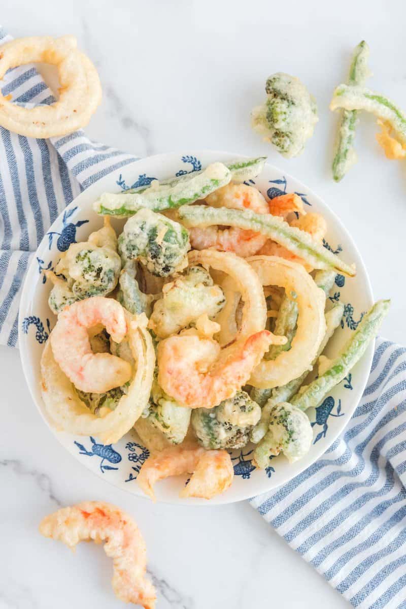 An overhead shot of a blue and white patterned bowl filled with a variety of shrimp and vegetable tempura. The light, crispy coating is visible on the shrimp, onion rings, broccoli, and green beans.