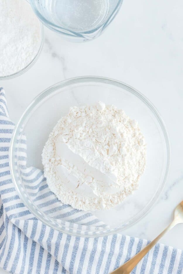 A clear glass bowl filled with the combined dry ingredients, all-purpose flour and cornstarch, for the tempura batter. The shot is taken from above, with a blue and white striped napkin to the side.