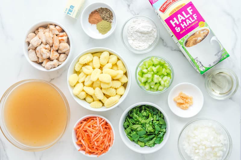 An overhead view of the ingredients for Olive Garden chicken gnocchi soup on a marble surface. Pictured are bowls of cooked chicken, gnocchi, chicken broth, carrots, celery, onion, spinach, and other components like half-and-half, butter, flour, and spices.