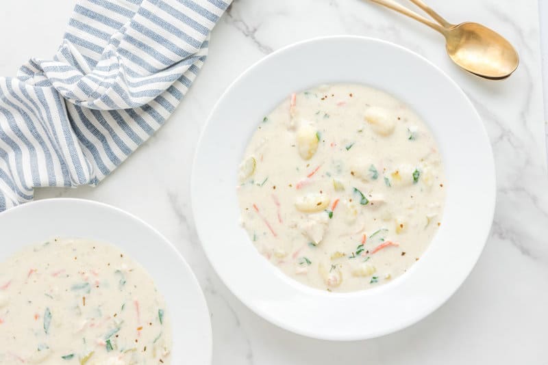 Two white bowls of creamy chicken gnocchi soup are served on a marble table. Each bowl is filled with the rich broth, tender gnocchi, chicken, and colorful vegetables. A striped napkin and gold spoons are placed nearby.