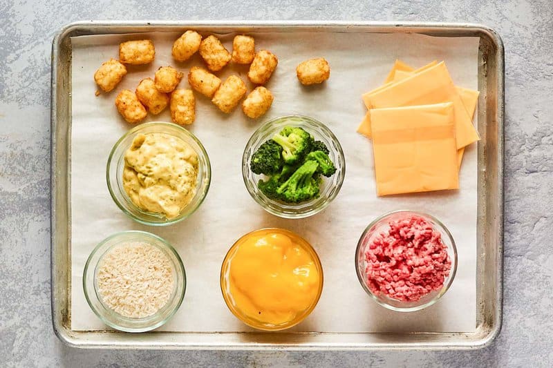An overhead view of the ingredients for broccoli tater tot casserole on a parchment-lined baking sheet. Pictured are raw ground beef, frozen broccoli florets, Velveeta cheese slices, frozen tater tots, and glass bowls containing cheddar cheese soup, broccoli cheese soup, and uncooked white rice.