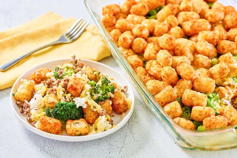 A serving of broccoli tater tot casserole on a white plate next to the full glass baking dish. The plated portion shows the mix of ground beef, broccoli, and rice, while the casserole dish highlights the crispy, golden-brown tater tot topping.