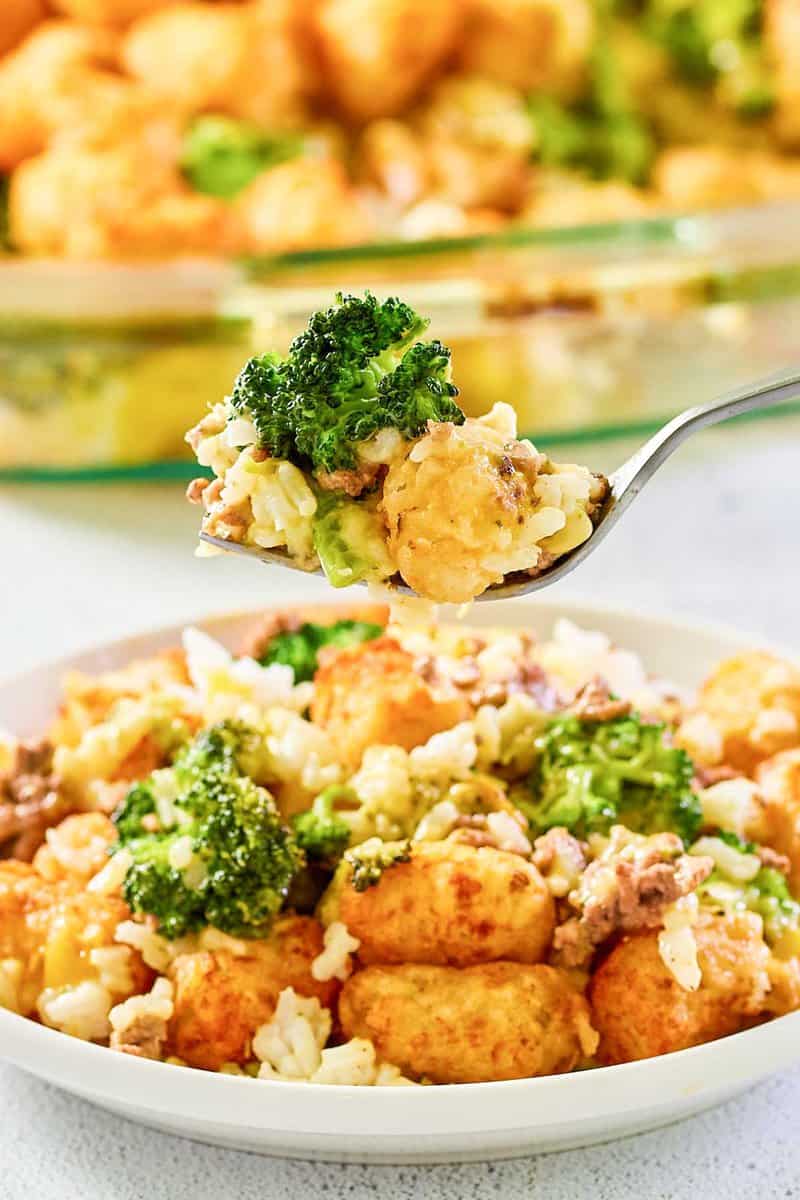 A close-up shot of a fork lifting a bite of broccoli tater tot casserole. The bite on the fork showcases the texture and ingredients, including a golden tater tot, a piece of broccoli, ground beef, and rice in a creamy sauce.