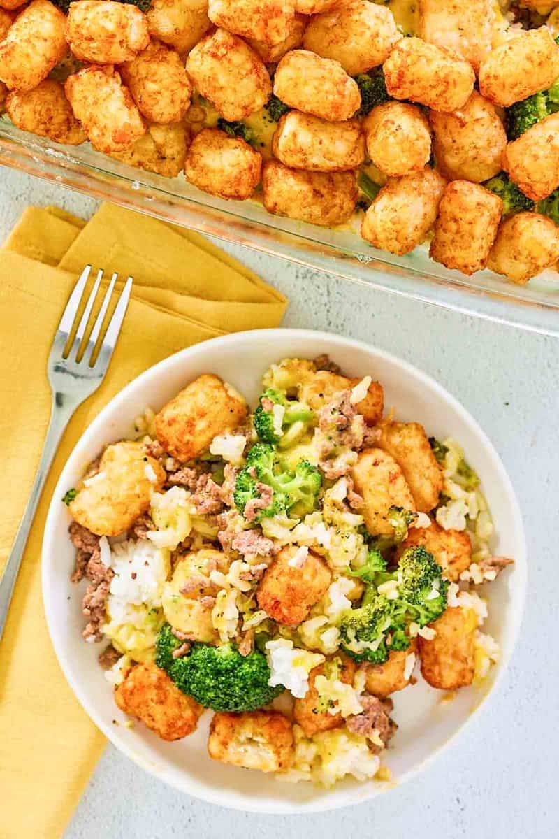 A serving of broccoli tater tot casserole on a white plate next to the full glass baking dish. The plated portion shows the mix of ground beef, broccoli, and rice, while the casserole dish highlights the crispy, golden-brown tater tot topping.