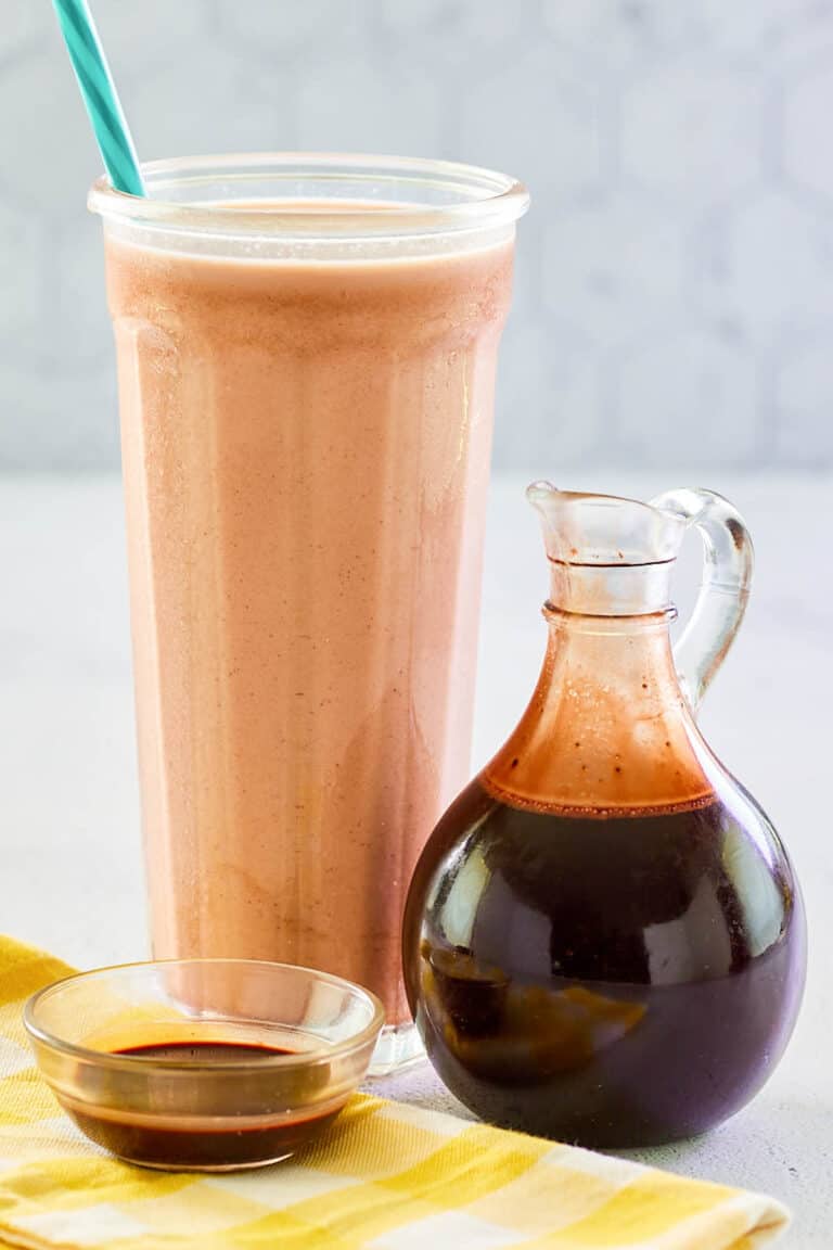 Homemade chocolate syrup in a small glass pitcher and a glass of chocolate milk.