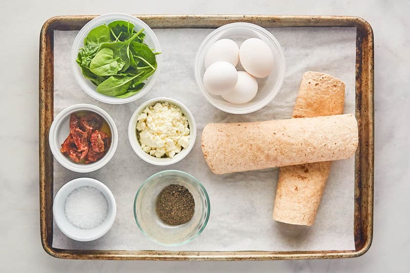n overhead view of the ingredients for a Starbucks spinach feta wrap on a baking sheet. Pictured are whole wheat tortillas, whole eggs, and bowls of fresh spinach, crumbled feta cheese, sun-dried tomatoes, salt, and pepper.