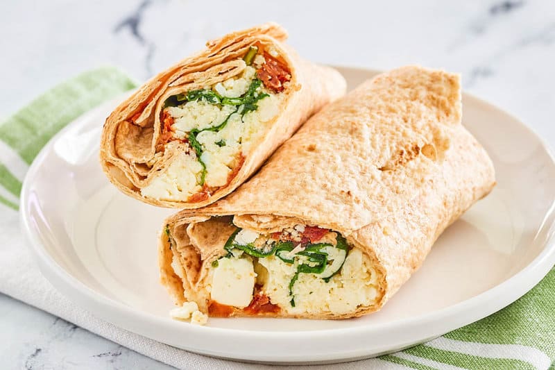 A homemade Starbucks spinach feta wrap, cut in half and stacked on a white plate. The cross-section clearly displays the filling of egg whites, spinach, sun-dried tomatoes, and feta cheese inside the whole wheat tortilla.