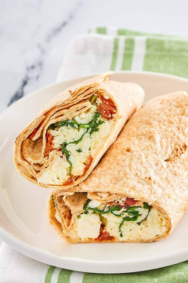 A close-up of a Starbucks-style spinach feta wrap cut in half. The image highlights the colorful filling of fluffy egg whites, green spinach, and red sun-dried tomatoes wrapped in a whole wheat tortilla.