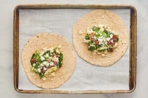 Two whole wheat tortillas are laid flat on a baking sheet, each with a scoop of the spinach, egg white, sun-dried tomato, and feta filling in the center, ready to be folded into wraps
