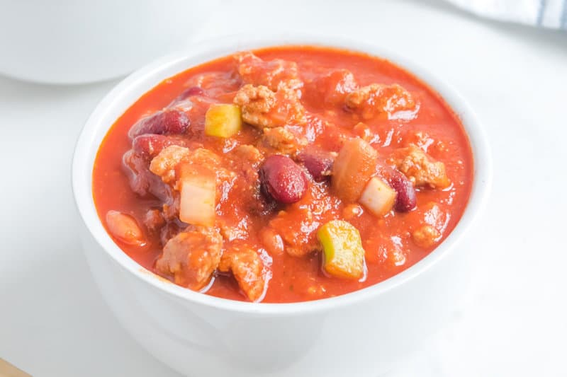 A close-up shot of a white bowl filled with a serving of copycat Wendy's chili. The thick, hearty texture is visible, showing ground beef, kidney beans, pinto beans, and diced vegetables in a rich tomato sauce.