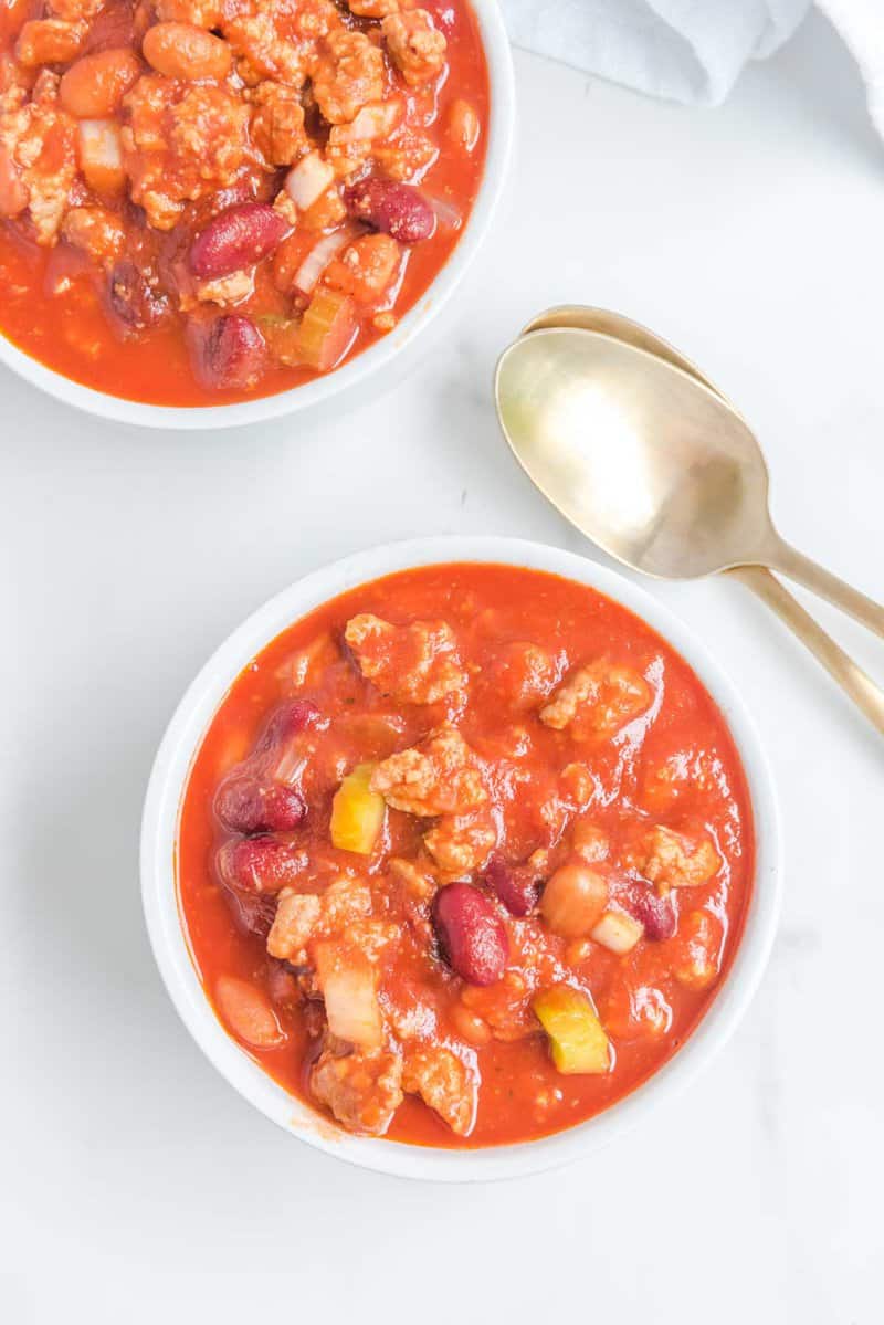 An overhead view of two white bowls filled with copycat Wendy's chili, ready to be eaten. The bowls are arranged on a white surface with two gold spoons beside them.