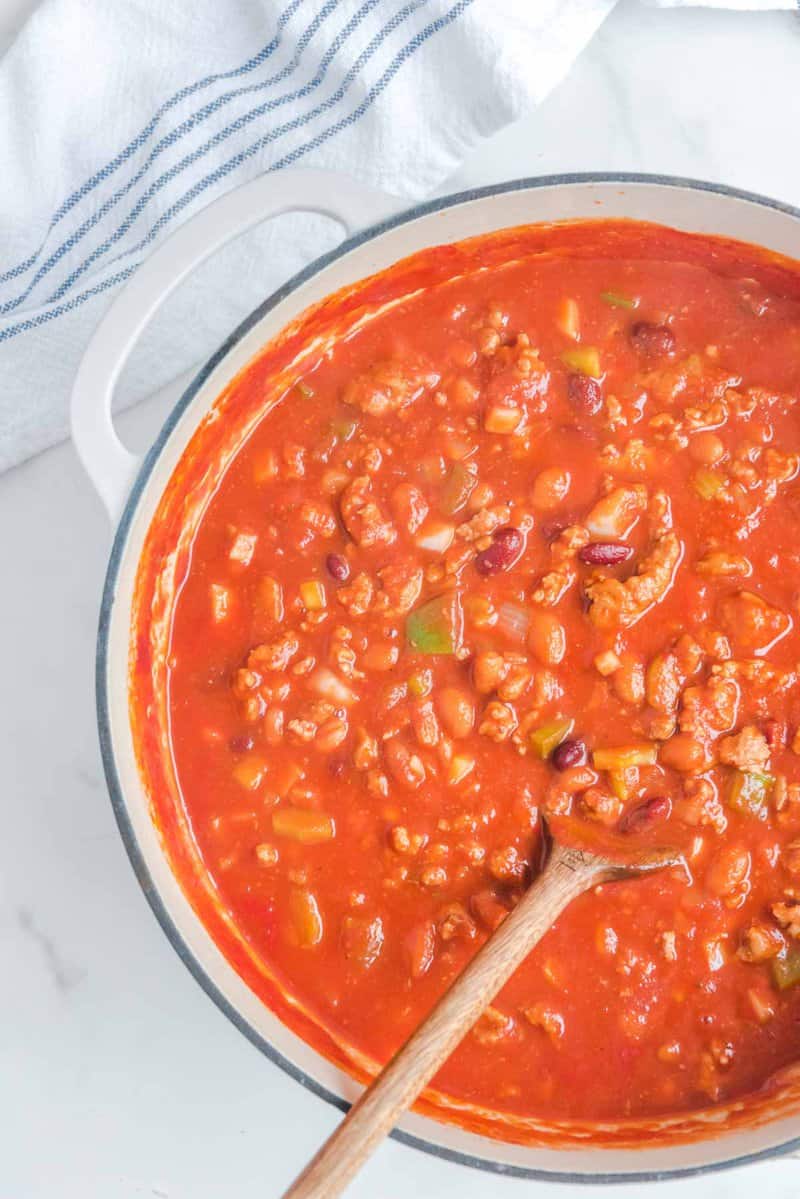 n overhead view of a large white pot filled with rich, hearty Wendy's chili. A wooden spoon stirs the chili, showing its thick texture with ground beef, beans, and vegetables.