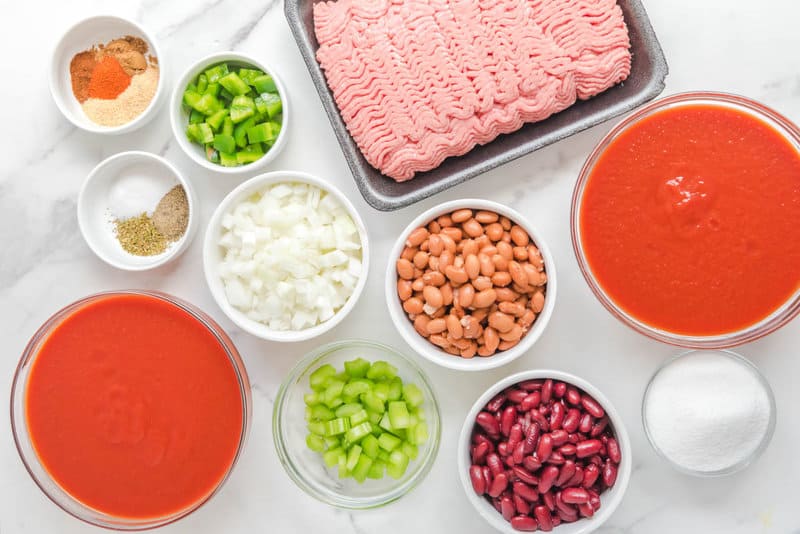 An overhead view of the ingredients for Wendy's chili on a marble countertop. Pictured are raw ground beef and bowls containing diced onion, celery, green bell pepper, kidney beans, pinto beans, tomato juice, tomato puree, and various spices.