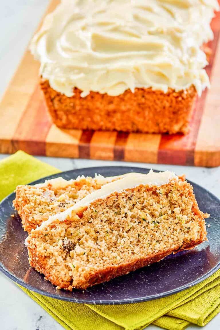 Two slices of zucchini nut bread with cream cheese frosting on a plate with a loaf behind it.