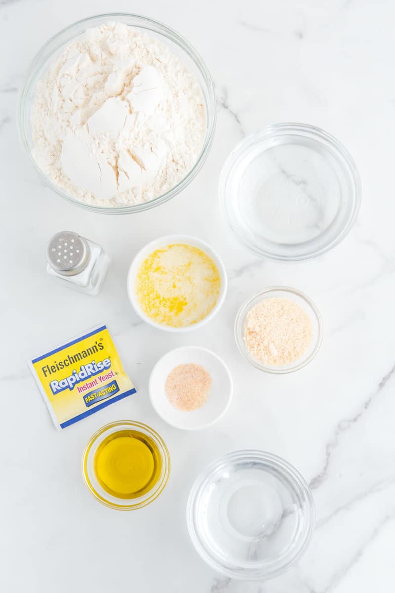 Flour, water, melted butter, oil, parmesan, garlic salt, and yeast packet on a white counter.