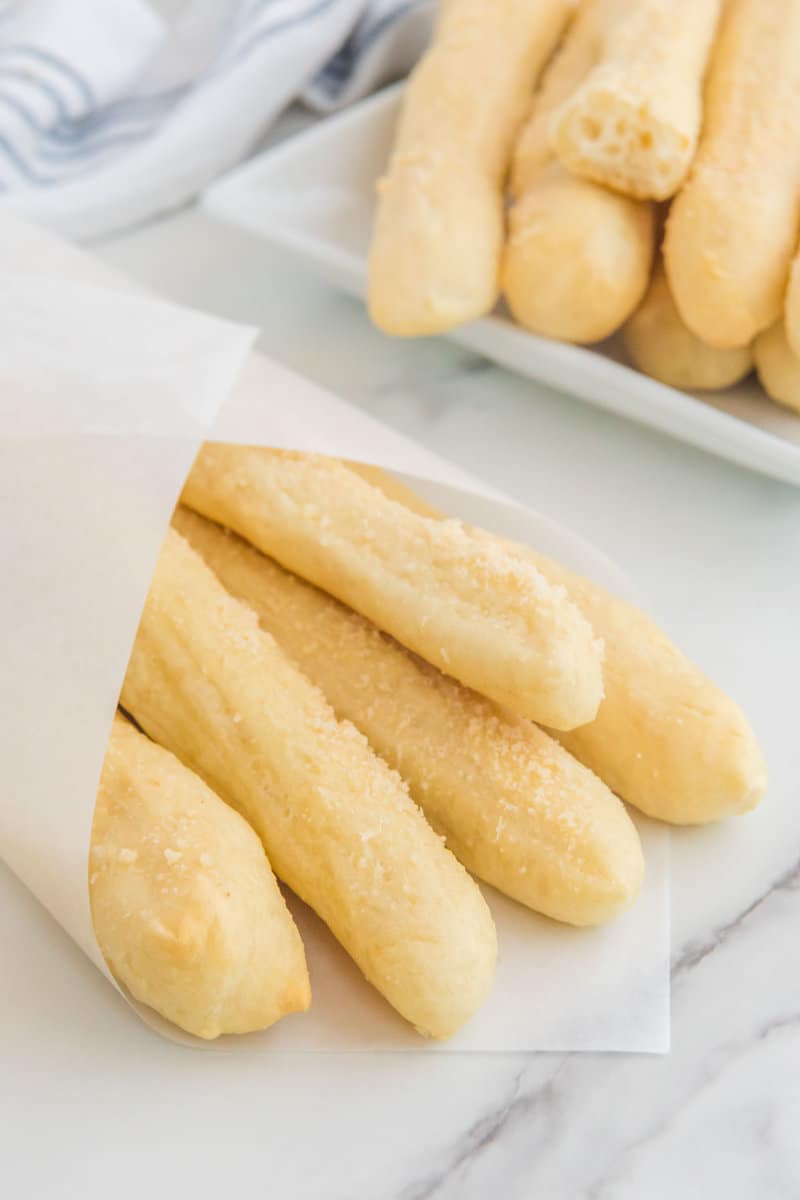 Golden brown homemade Crazy Bread sticks in a white paper sleeve with more on a plate behind.