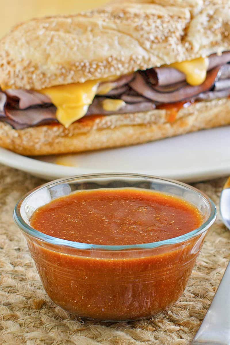 A glass bowl of red Arby's sauce sits in the foreground, with a roast beef and cheddar sandwich on a plate behind it.