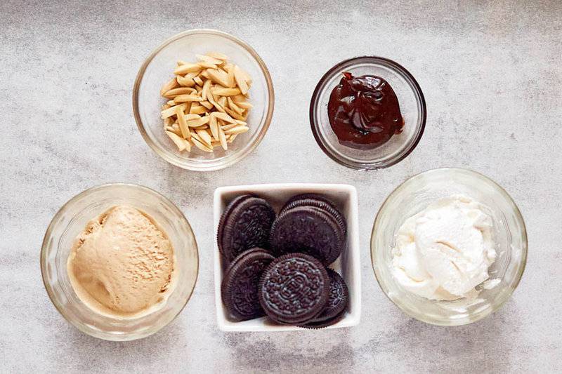 An overhead view of the ingredients for a no-bake mud pie. Pictured are bowls containing coffee ice cream, whipped cream, hot fudge, and slivered almonds, along with a central bowl of Oreo cookies for the crust.