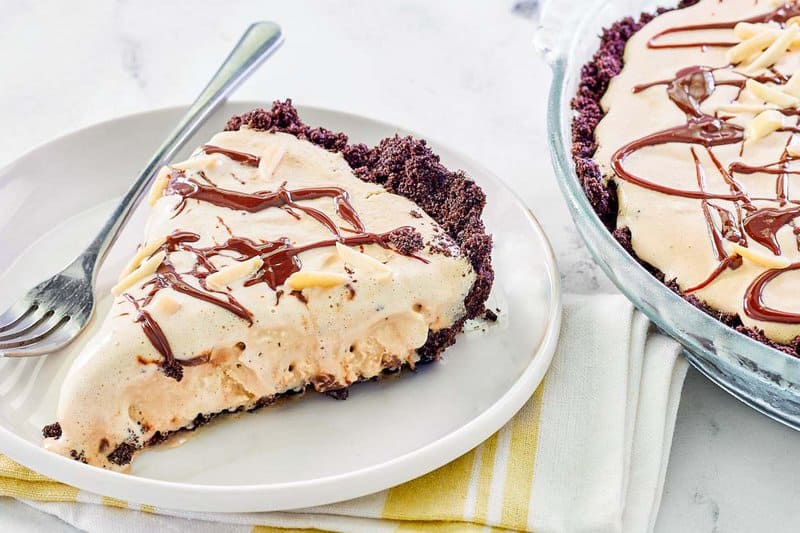 A slice of frozen mud pie is served on a white plate, with the rest of the pie visible in the background. The slice features a thick Oreo cookie crust, a coffee ice cream filling, a chocolate drizzle, and an almond topping.