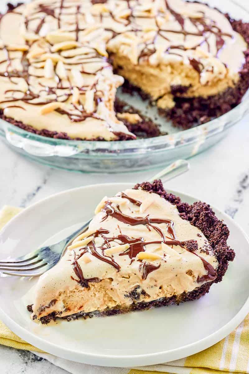 A slice of frozen mud pie is served on a white plate, with the rest of the pie visible in the background. The slice features a thick Oreo cookie crust, a coffee ice cream filling, a chocolate drizzle, and an almond topping.