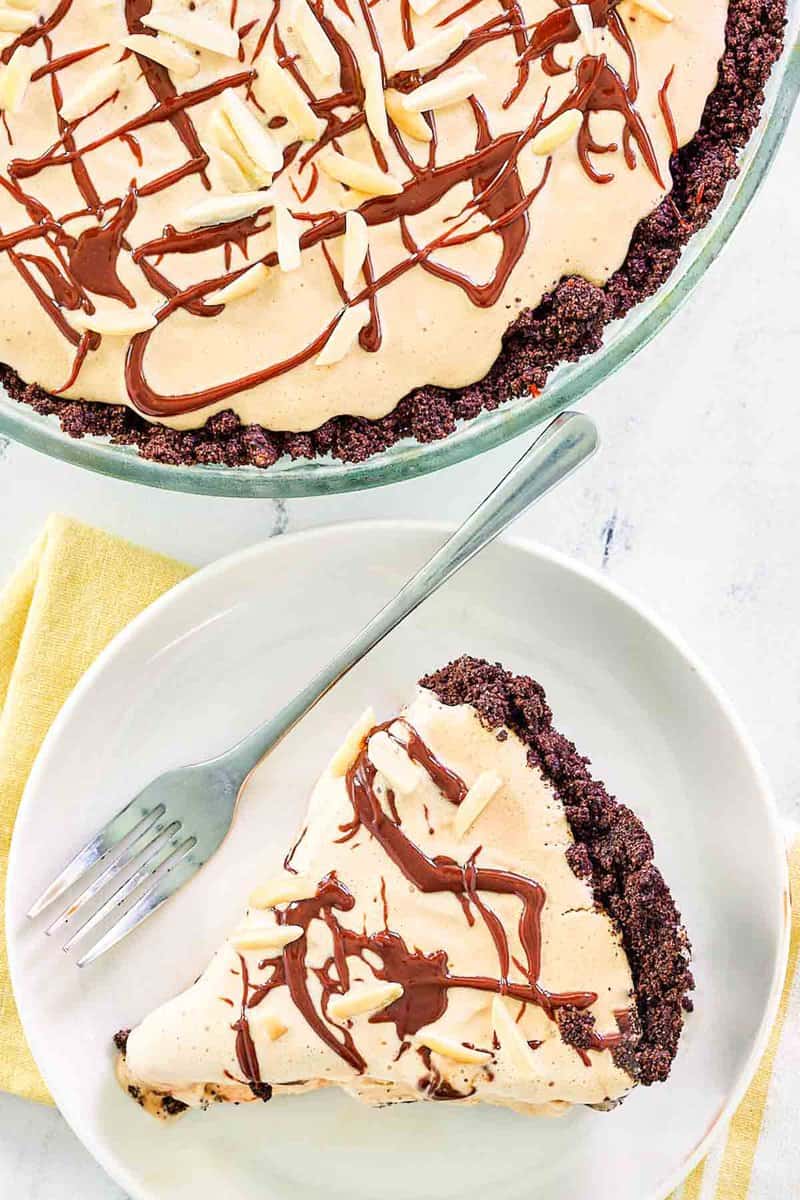 An overhead, flat-lay shot of a slice of mud pie on a plate next to the full pie. Both the slice and the pie showcase a thick Oreo crust, a coffee ice cream filling, and a topping of chocolate drizzle and slivered almonds.