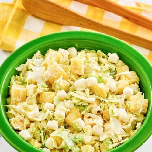 A bowl of pineapple coleslaw and salad tongs behind it.
