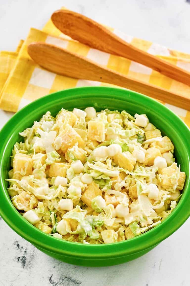 A bowl of pineapple coleslaw and salad tongs behind it.