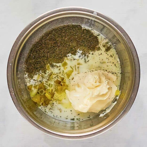 An overhead shot of a stainless steel mixing bowl with all the dill pickle ranch dressing ingredients inside before being whisked. Mayonnaise, buttermilk, chopped pickles, and dried herbs are shown layered together.