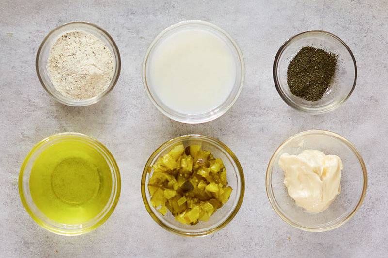 An overhead view of the six ingredients for dill pickle ranch dressing arranged in small glass bowls. Pictured are bowls of dry ranch seasoning mix, buttermilk, dried dill, pickle juice, chopped dill pickles, and mayonnaise.