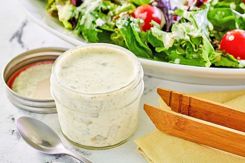 A close-up shot of the finished dill pickle ranch dressing in a small glass jar, highlighting its creamy texture and visible pieces of pickle and dill. A fresh green salad sits in the blurred background, suggesting a perfect pairing.