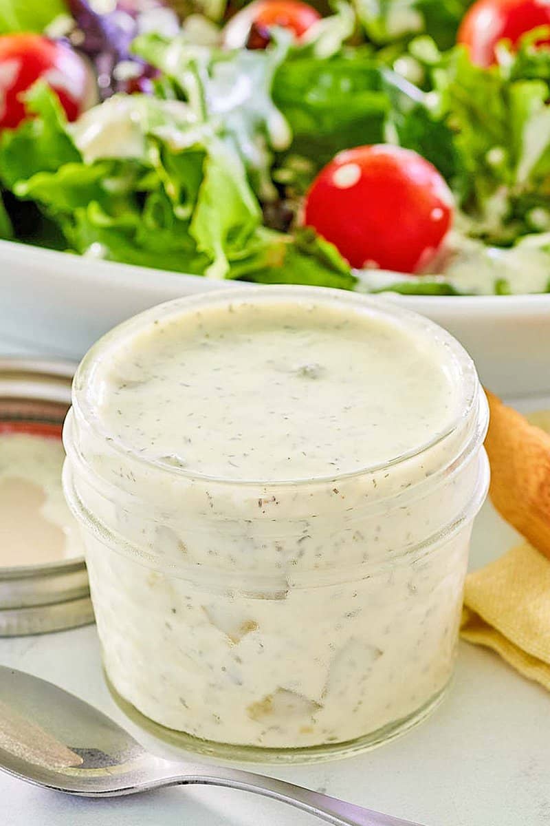 An overhead, flat-lay photo of dill pickle ranch dressing. A small glass bowl of the creamy dressing is in the foreground, while a fresh salad drizzled with the dressing is partially visible on a plate in the background.