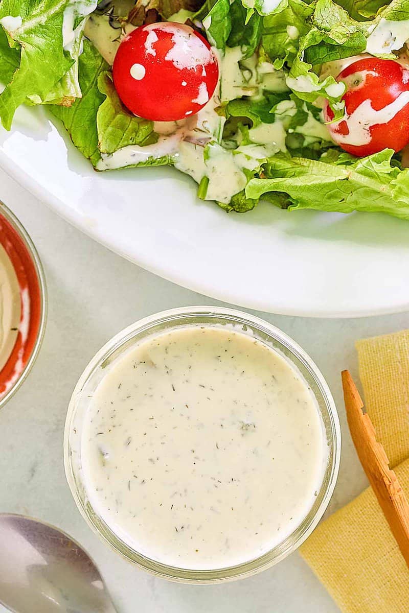 An overhead, flat-lay photo of dill pickle ranch dressing. A small glass bowl of the creamy dressing is in the foreground, while a fresh salad drizzled with the dressing is partially visible on a plate in the background.